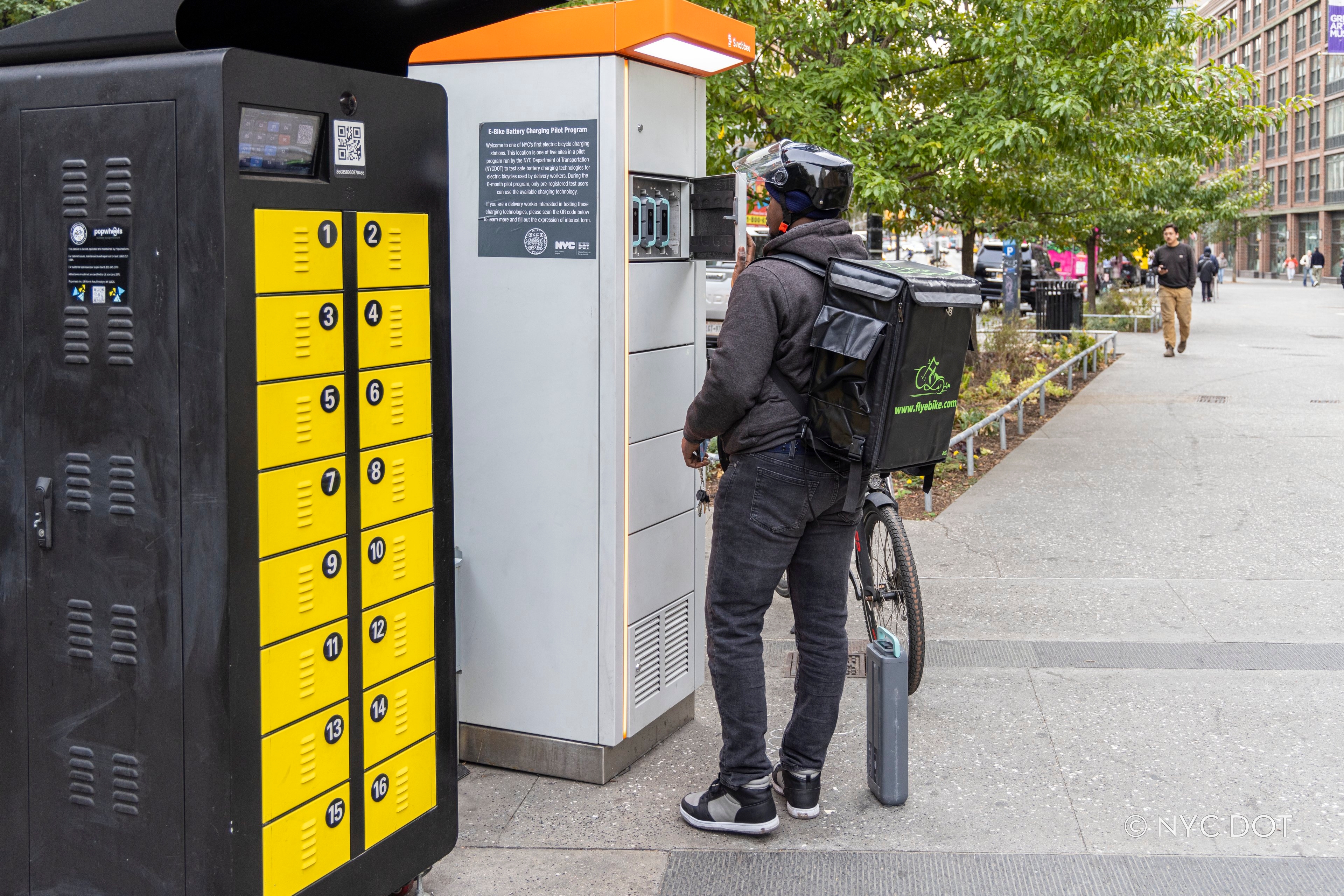 A delivery worker wearing a helmet and a green insulated delivery backpack stands at an outdoor e-bike battery swapping cabinet on a city sidewalk. A white truck and urban buildings are visible in the background.