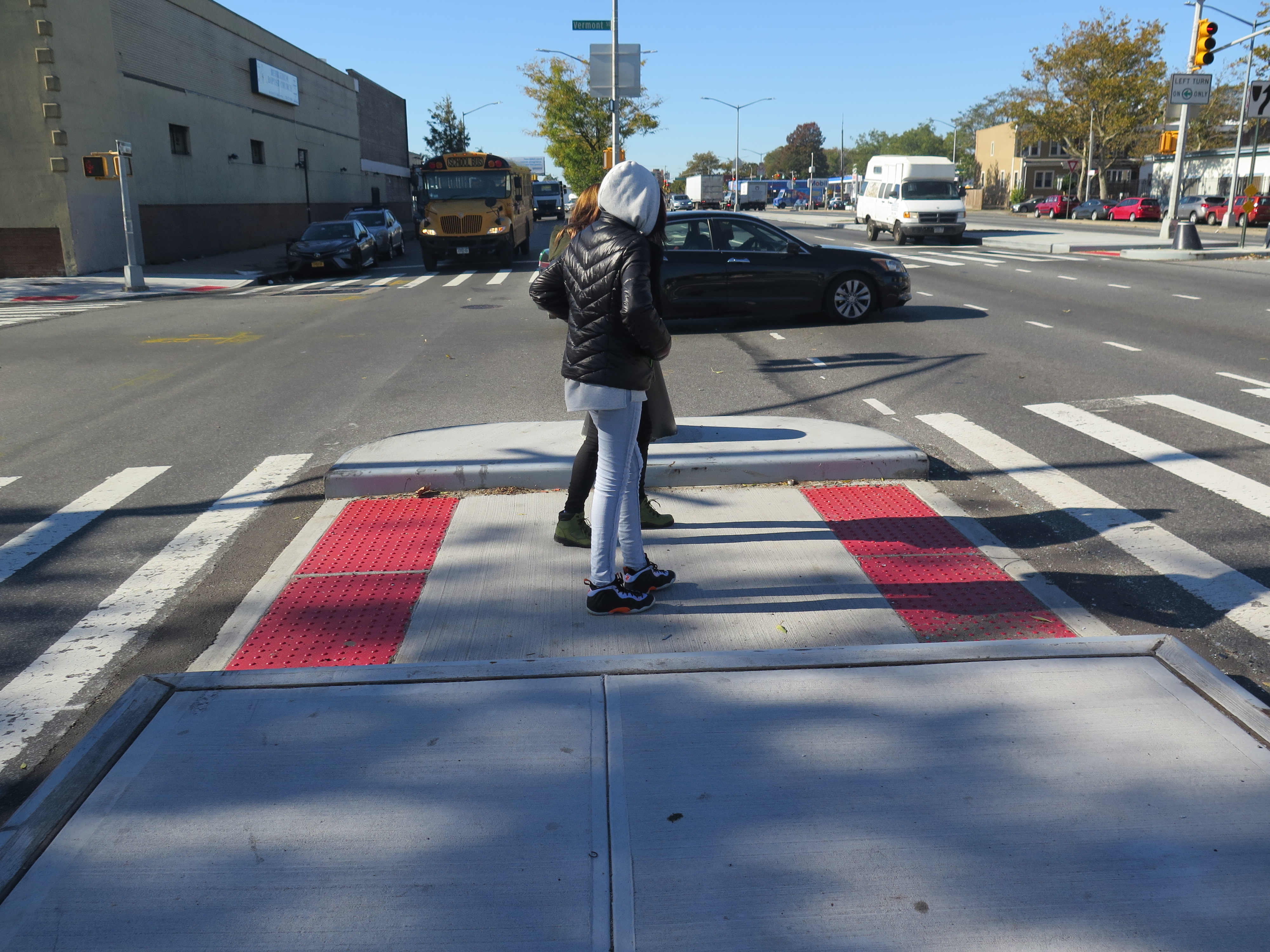 Two pedestrians crossing Linden Blvd at Vermont St, pausing in a  newly constructed median tip 