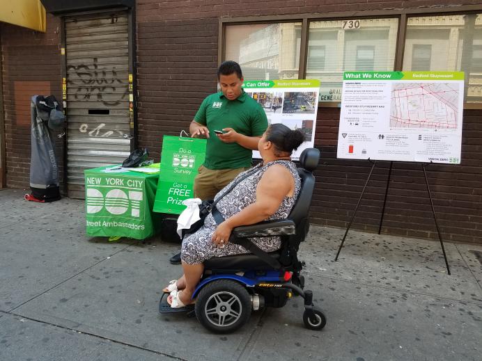 An Ambassador speaks with a person in a motorized wheelchair outside of the Broadway and Flushing JMZ stop.