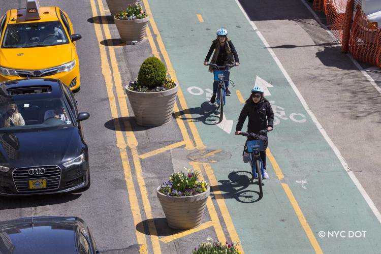 Two cyclists riding Citi Bikes in a bike lane pass by a row of cars stuck in traffic. 