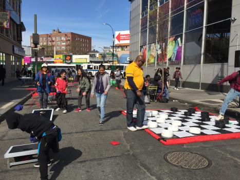 A scene from the One Day Plaza. In it, a group of children can be seen playing Corn Hole, while there are two adults playing a giant version of checkers. 