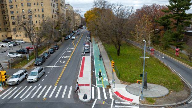 On Webster Ave in the Bronx, a cyclist and e-scooter use a separated greenway while a pedestrian crosses at a new concrete island and bus stop.