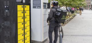 A delivery worker wearing a helmet and a green insulated delivery backpack stands at an outdoor e-bike battery swapping cabinet on a city sidewalk. A white truck and urban buildings are visible in the background.